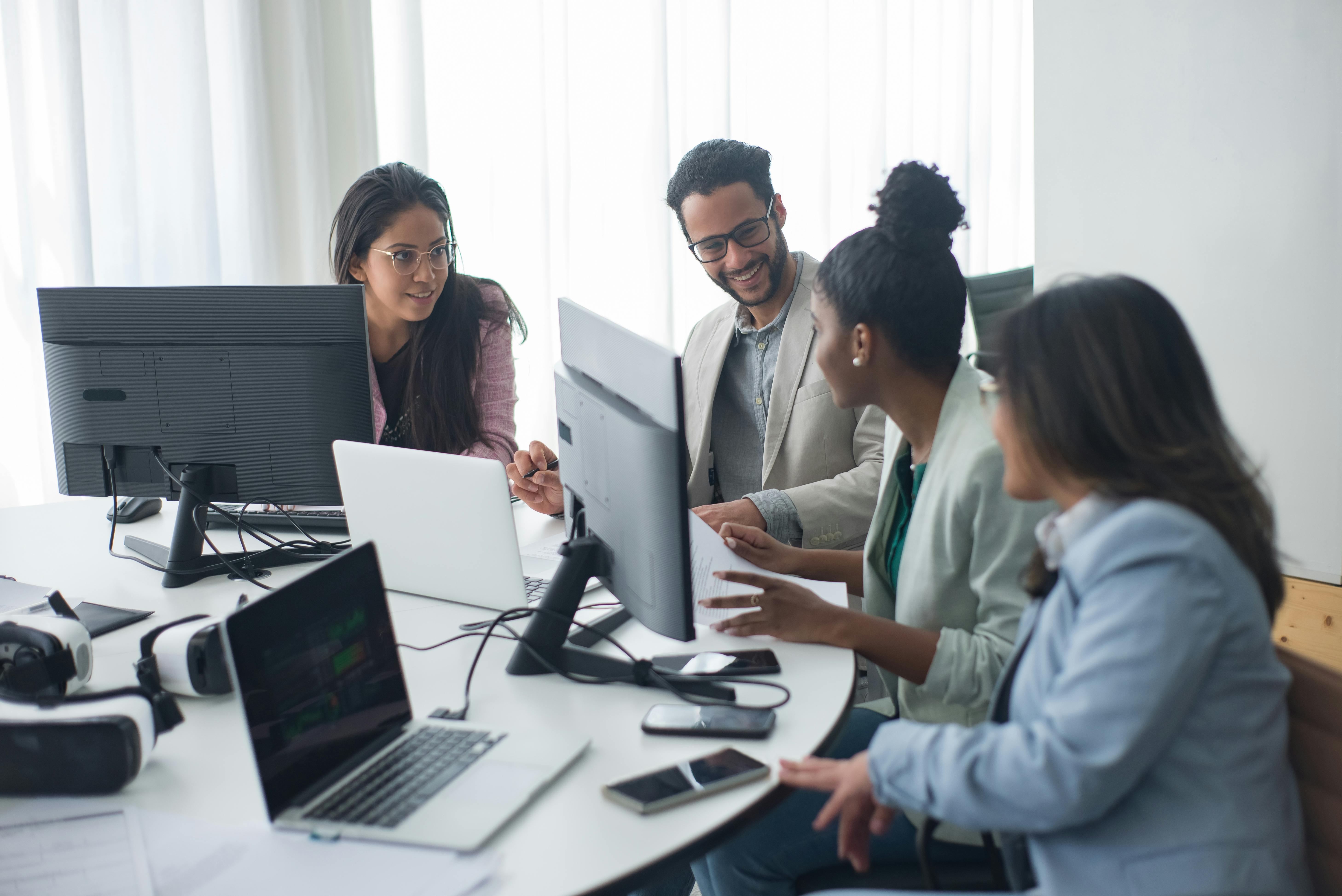 A group of colleagues sitting around a table in a modern office, engaged in a collaborative meeting with laptops and documents.