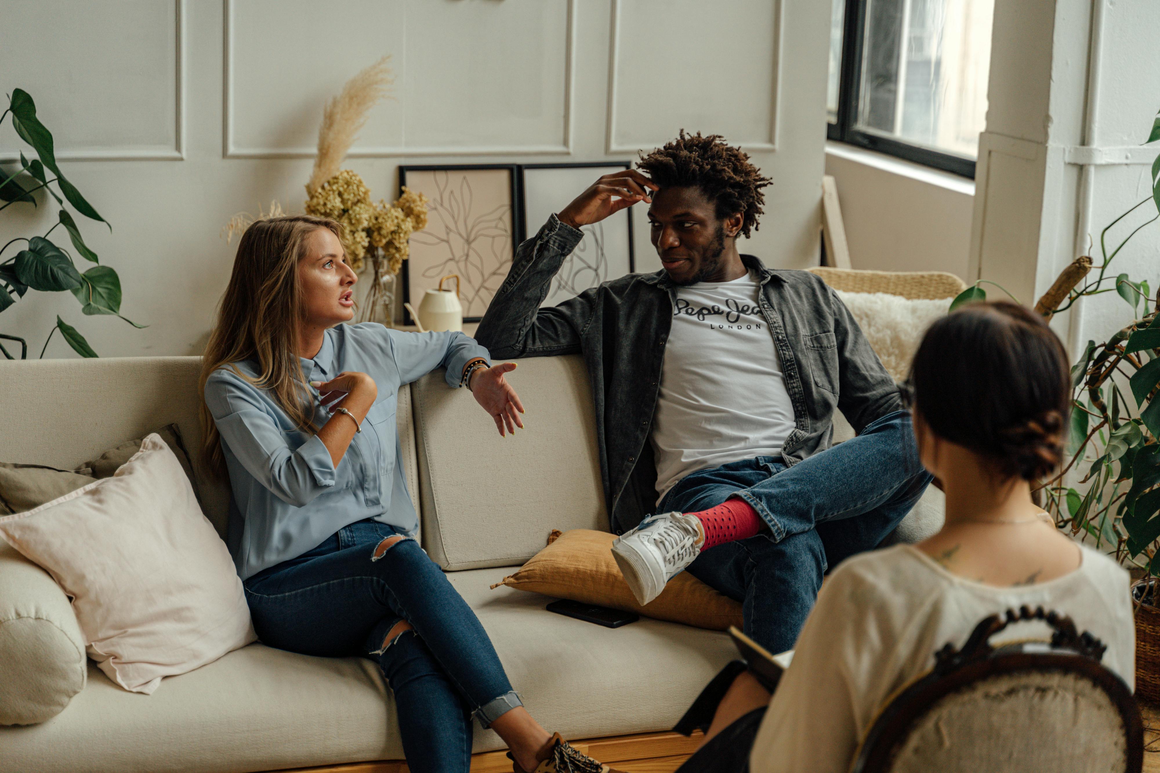 A group of three people sitting in a cozy, sunlit room having a thoughtful conversation. One woman is speaking with expressive hand gestures, while the others listen attentively, creating a sense of empathy and engagement.