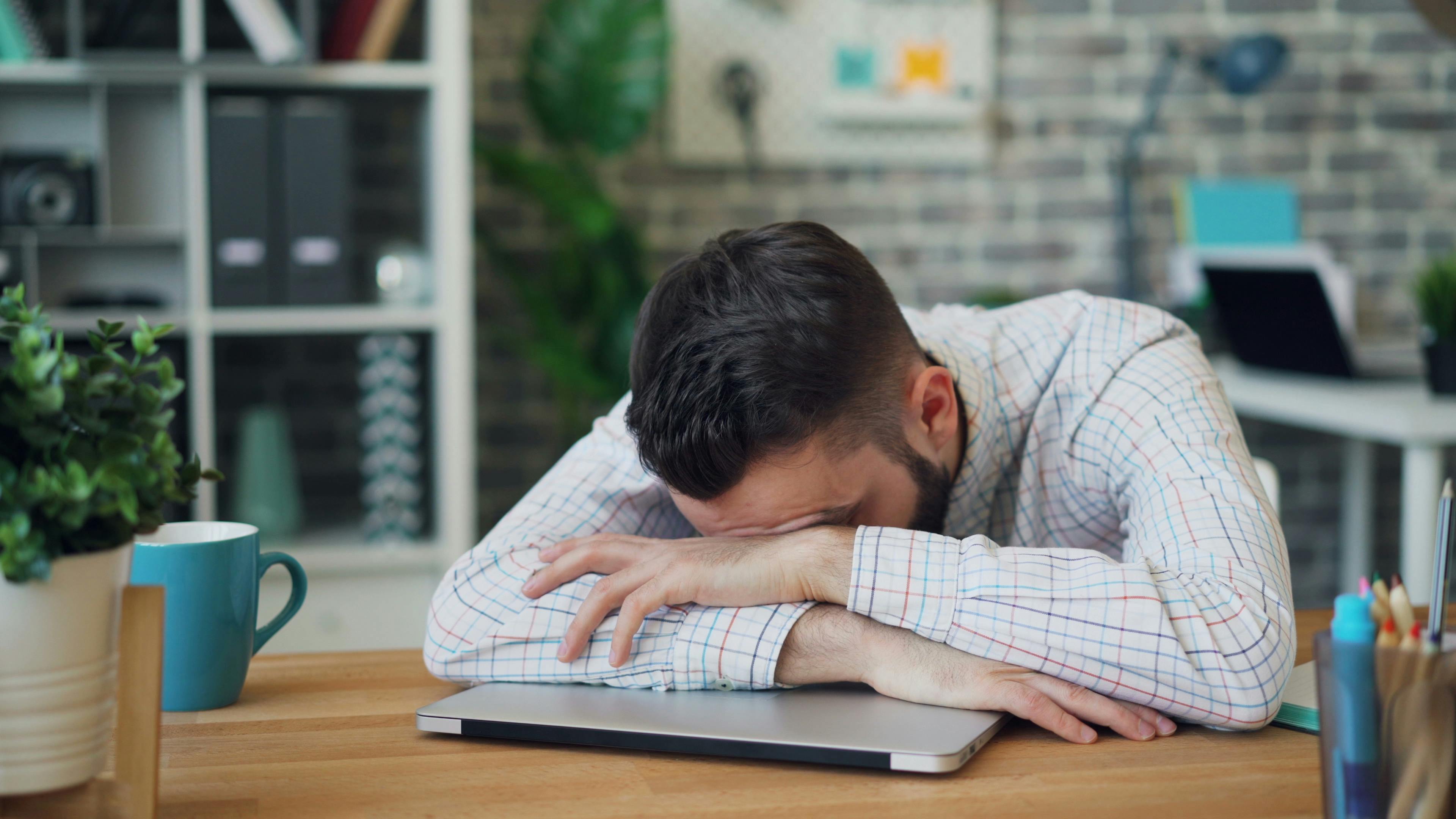 A man in a shirt lying face down on a table in an office, symbolizing workplace burnout and exhaustion.