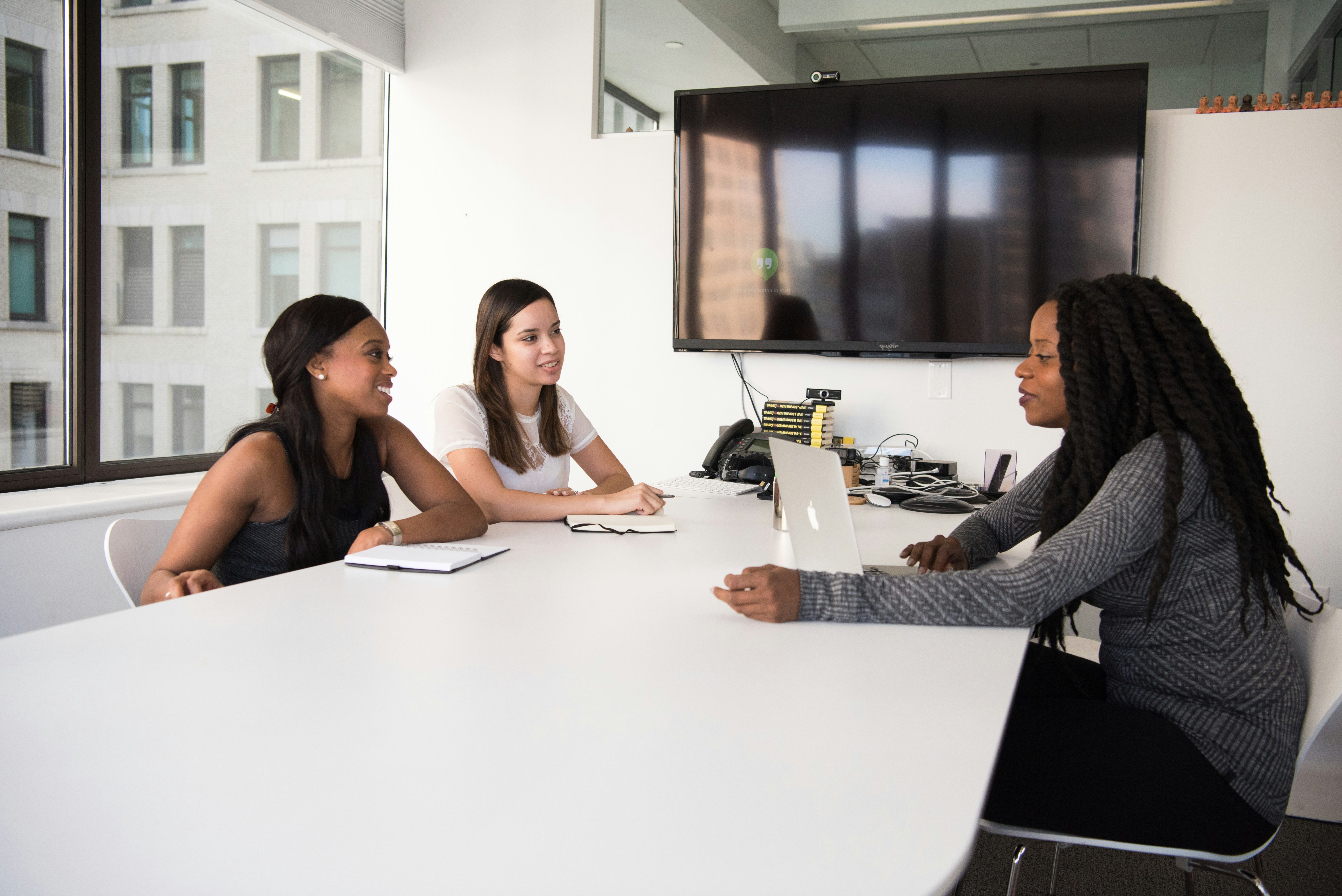 Three women in a modern office sitting around a white conference table, engaged in a collaborative meeting. One woman is using a laptop while the others listen attentively and take notes. A large monitor and office equipment are visible in the background.