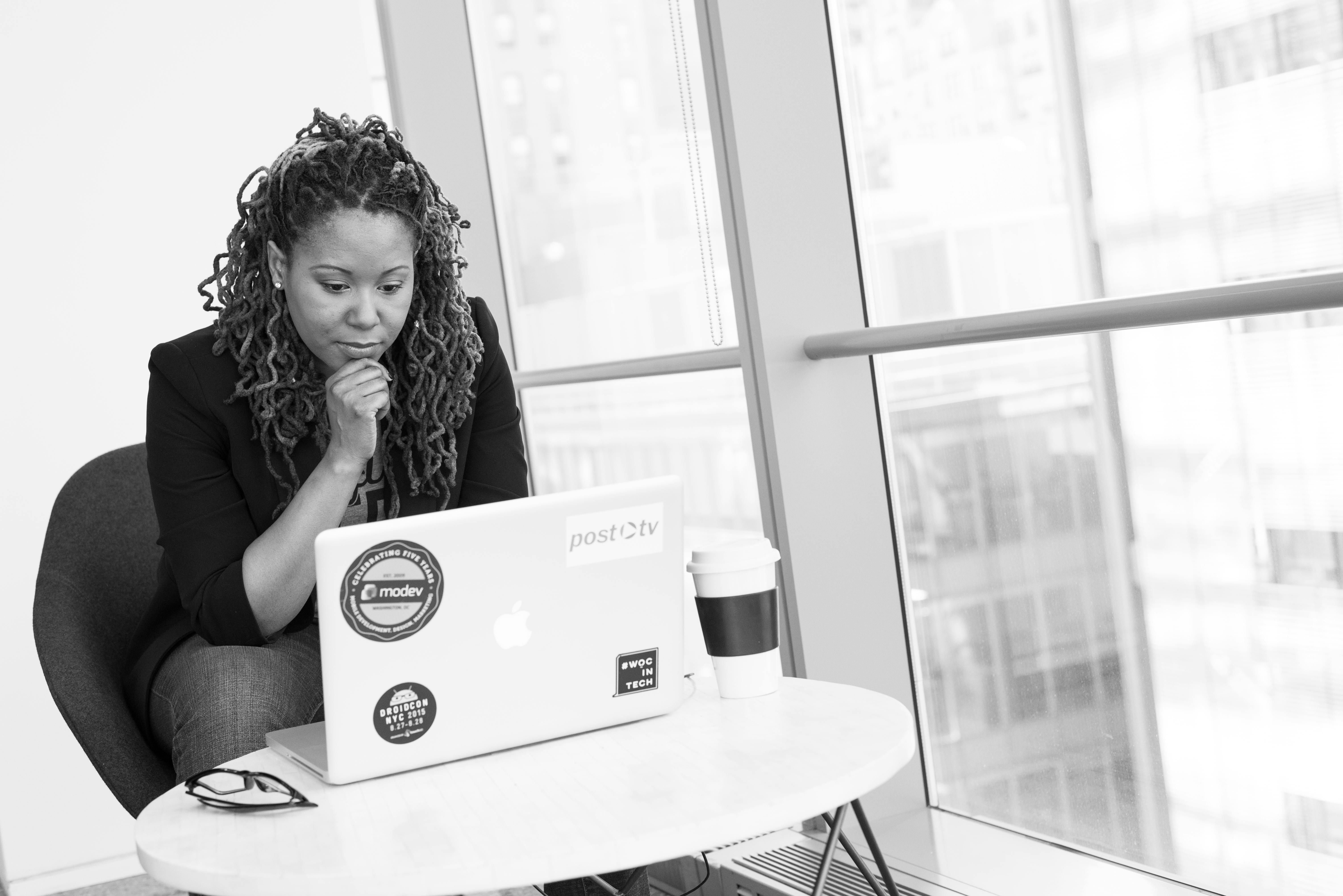 Grayscale photo of a woman sitting at a desk, focused on her MacBook, symbolizing strategic thinking and leadership in a professional environment.