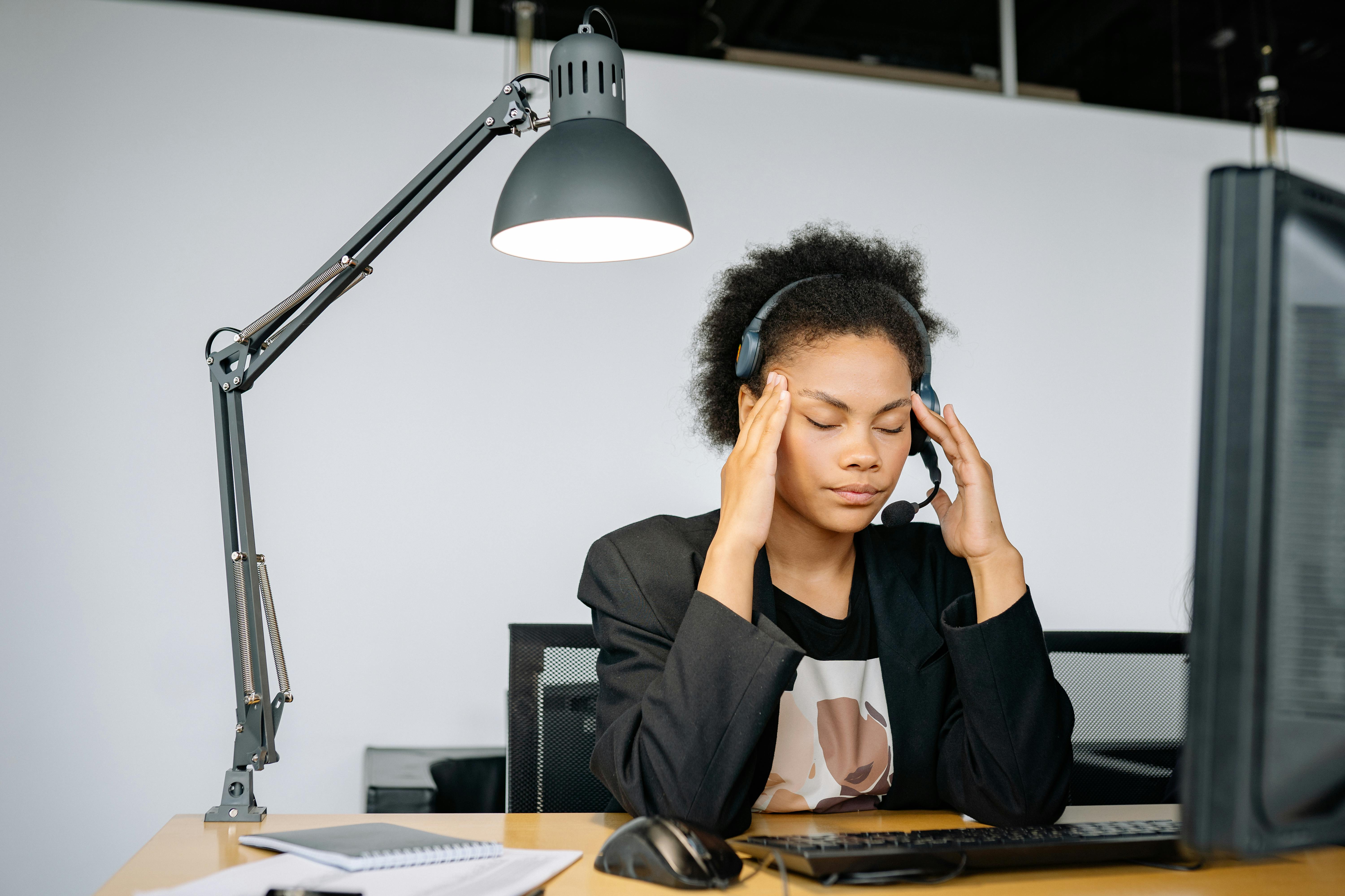 a young female contact center agent sitting at her desk with her headset on, rubbing her temples, eyes closed