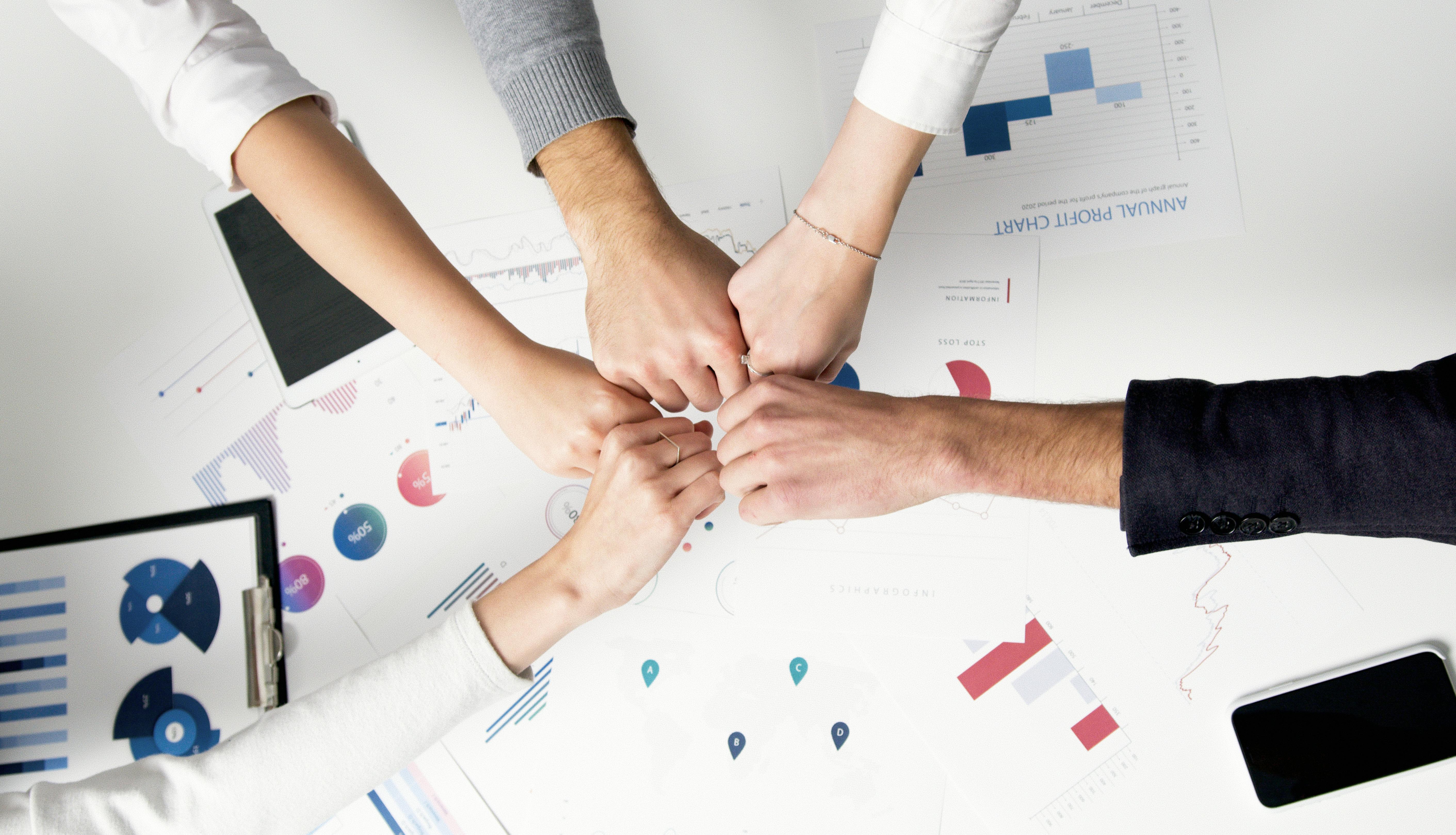an image of 5 hands in the middle of a table over some reports, signifying team work