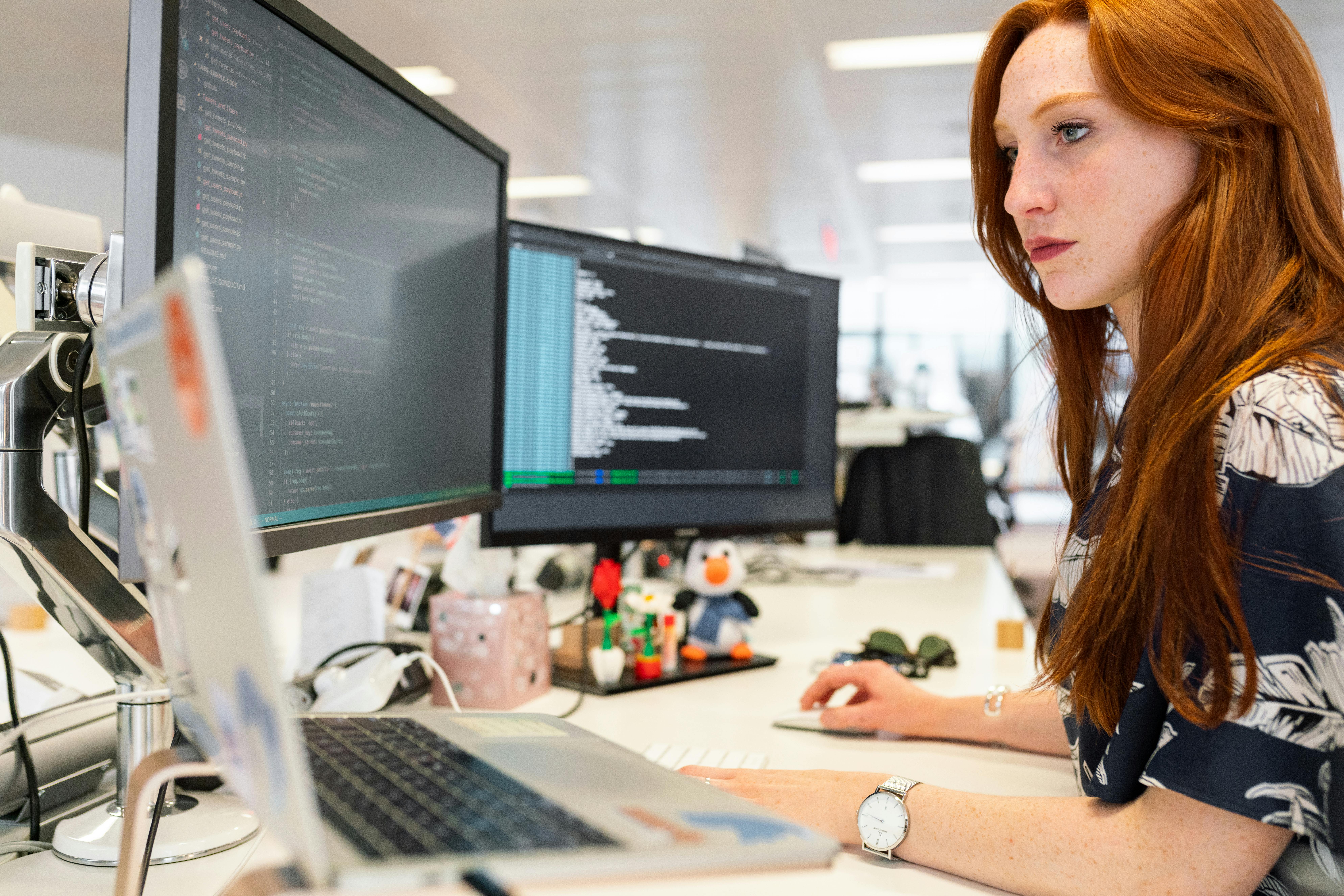 red headed woman sitting at desk at work with two computer screens